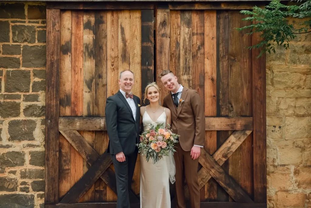 Bride in white gown holding bouquet, flanked by two suited men, standing before rustic wooden barn doors.