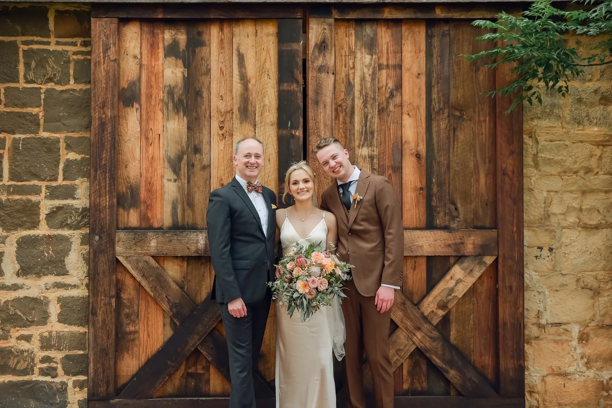Bride in white gown holding bouquet, flanked by two suited men, standing before rustic wooden barn doors.