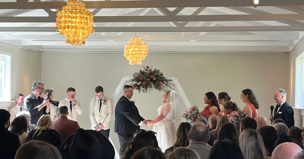 Bride and groom unite under a floral hexagon arch inside a high-beamed room with guests and party around them