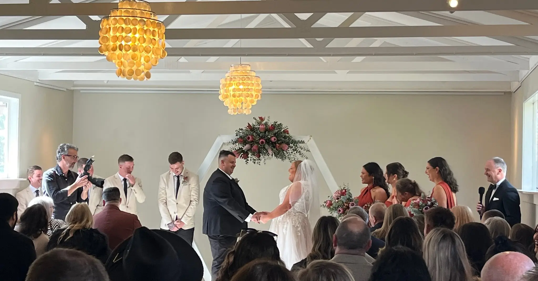 Bride and groom unite under a floral hexagon arch inside a high-beamed room with guests and party around them