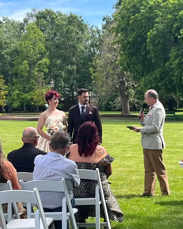 Bride and groom stand beneath floral arch exchanging vows, officiant behind in outdoor garden setting.