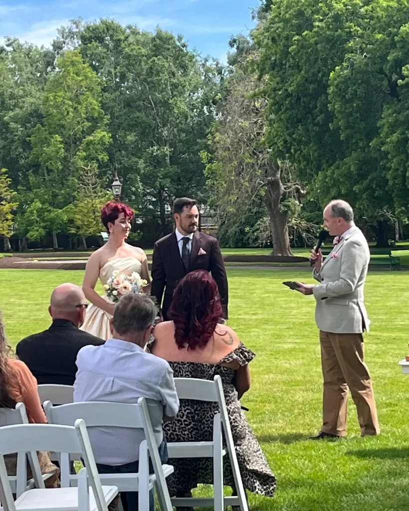 Bride and groom stand beneath floral arch exchanging vows, officiant behind in outdoor garden setting.