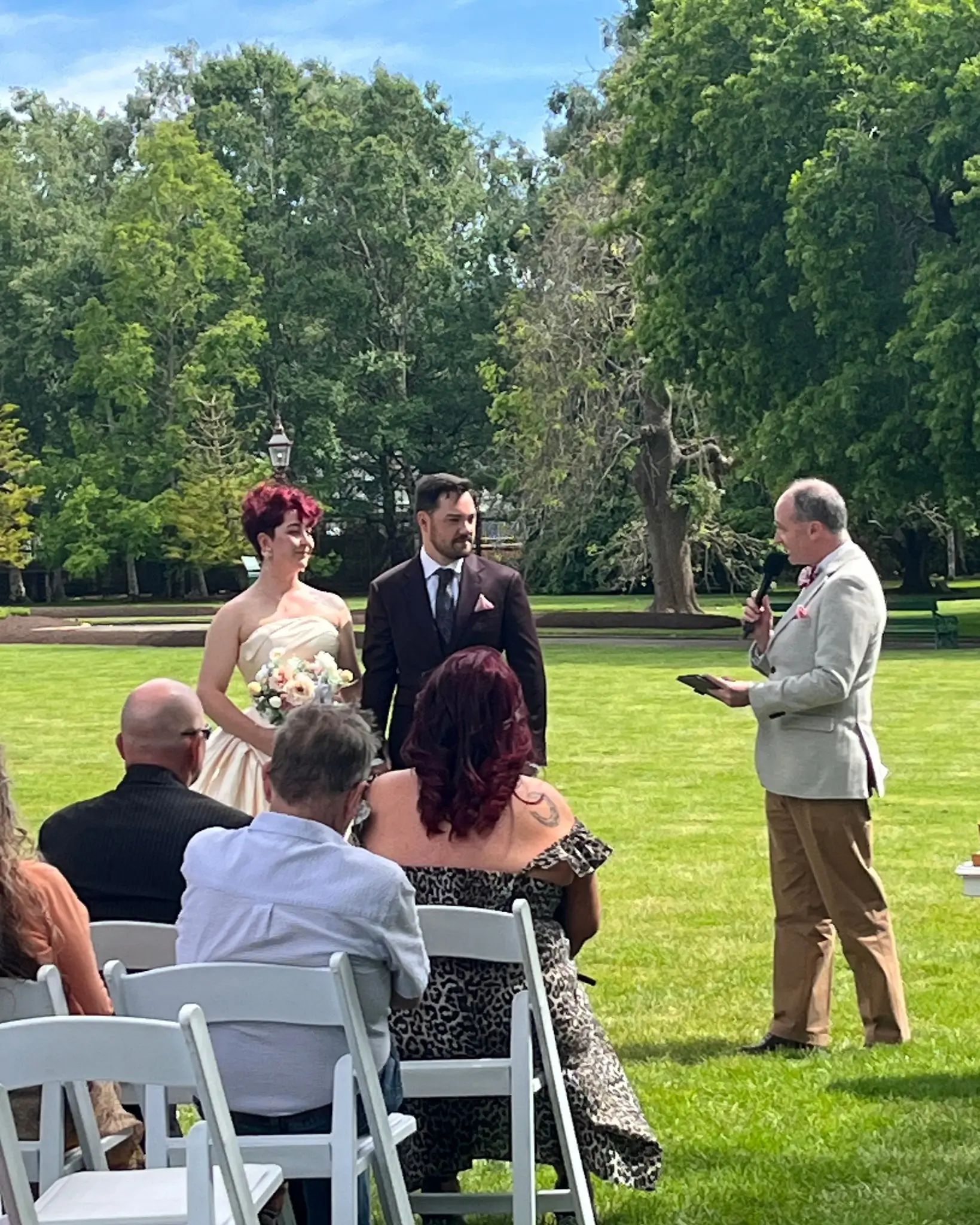 Bride and groom stand beneath floral arch exchanging vows, officiant behind in outdoor garden setting.