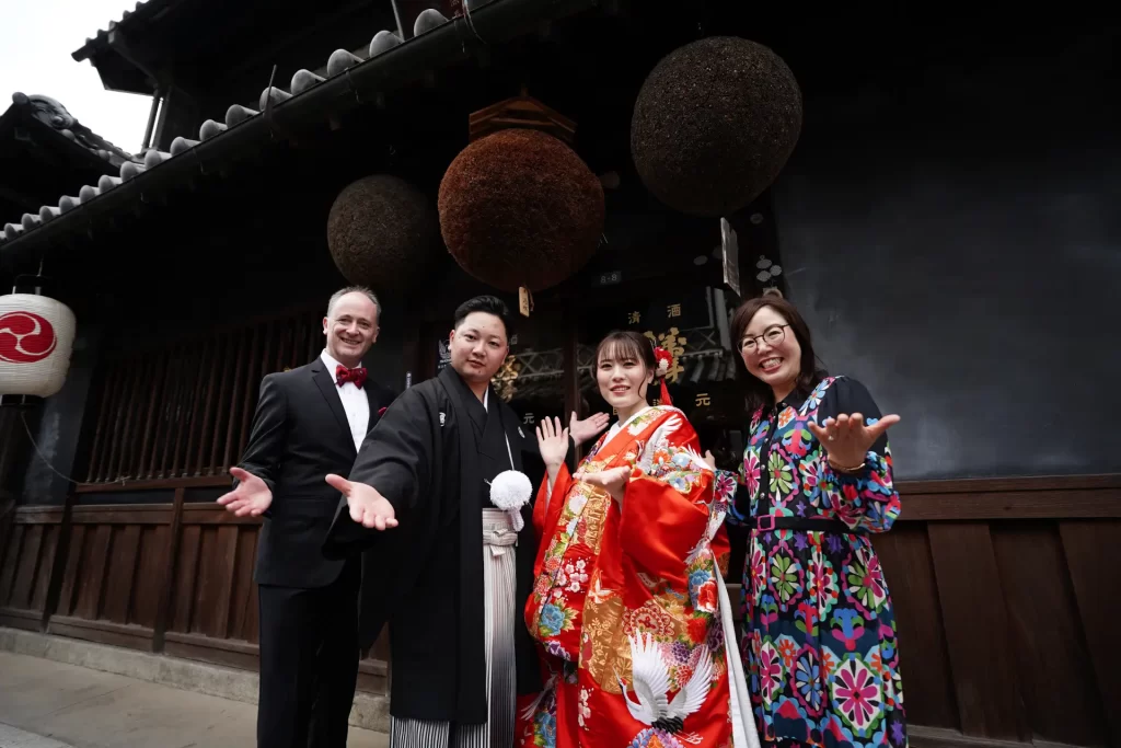 Bride in red kimono and groom in black robe pose with celebrant and guest outside traditional Japanese building