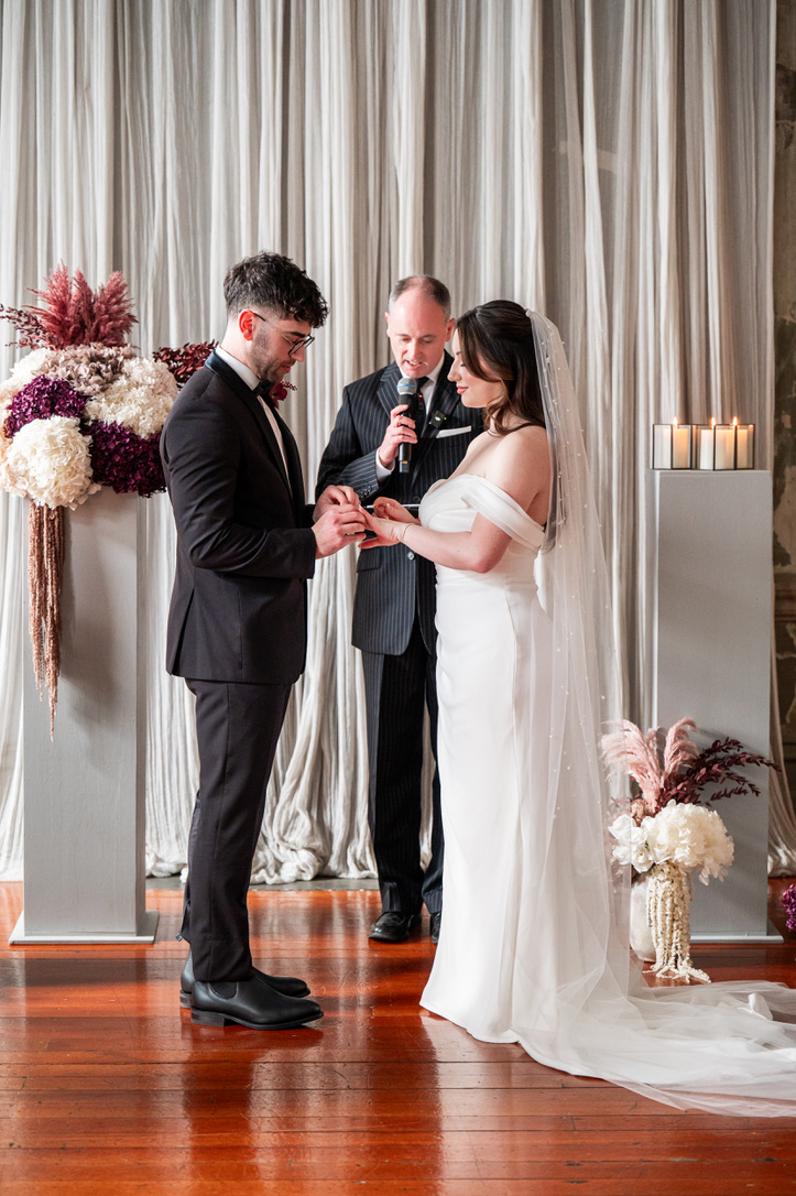 Bride and groom exchange rings during indoor ceremony, officiant speaking behind, floral pillars on each side.